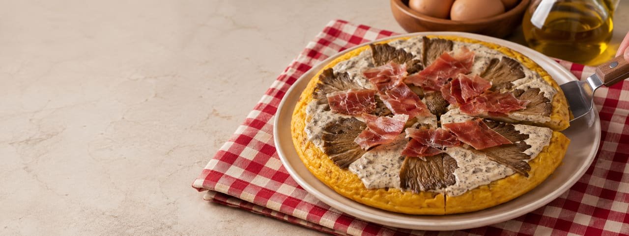 Tortilla de patata casera cortada en porciones, con una capa cremosa y jamón por encima, servida con pala sobre mantel de cuadros rojo y blanco; huevos y aceite de oliva de fondo